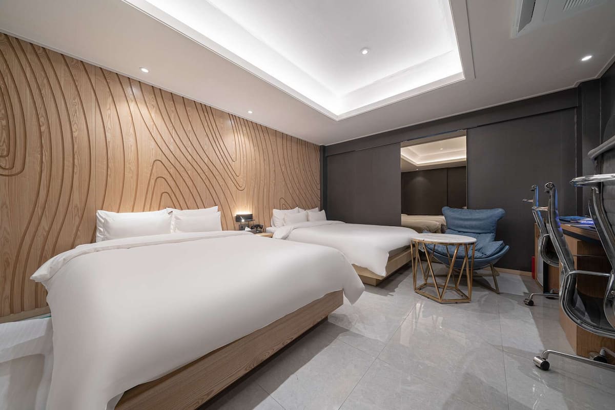 Une chambre d’hôtel moderne avec deux lits, une petite table, une chaise bleue et une coiffeuse avec un miroir.