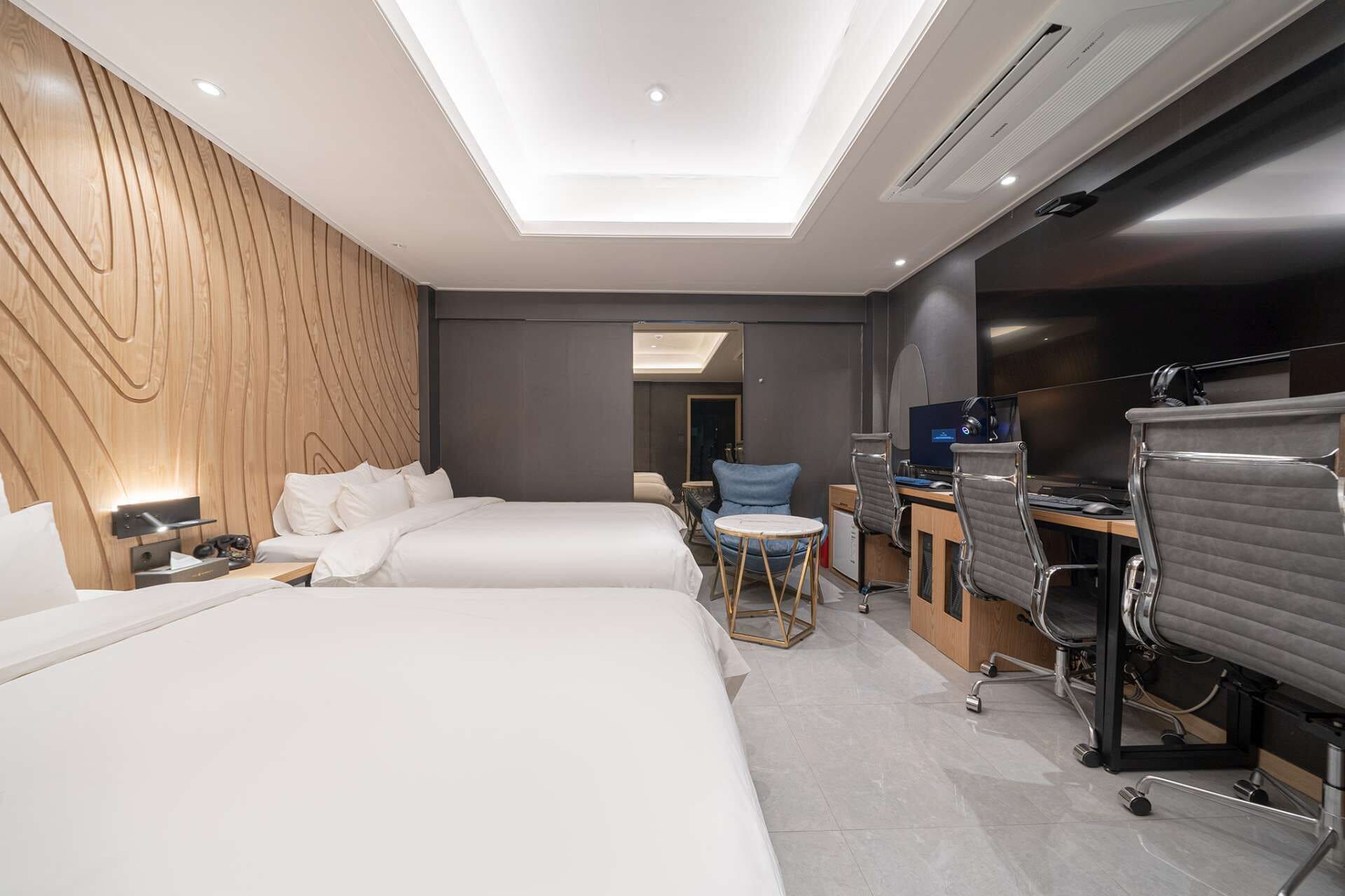 Una habitación de hotel moderna con dos camas, un escritorio con computadora y audífonos, una silla y un televisor grande.