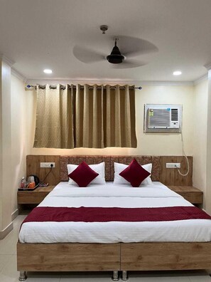 A hotel room with a bed, a ceiling fan, a window with curtains, an air conditioning unit, and a bedside table with a phone and a bottle. - Coco Inn Homestay  (Varanasi)