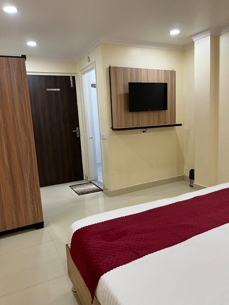 Een hotelkamer met een bed, een aan de muur bevestigde televisie, een houten garderobe en een deur naar een andere kamer.