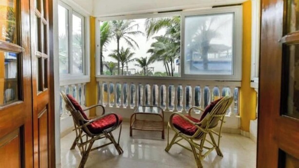 A room with two wicker chairs, a small table, and a view of palm trees through large windows. - Joy Guest House (Calangute)