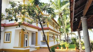 A two-story building with a red-tiled roof, yellow trim, and a covered patio area with palm trees. - Joy Guest House (Calangute)