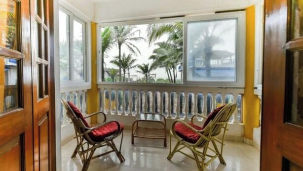 A room with two wicker chairs, a small table, and a view of palm trees through large windows. - Joy Guest House (Calangute)