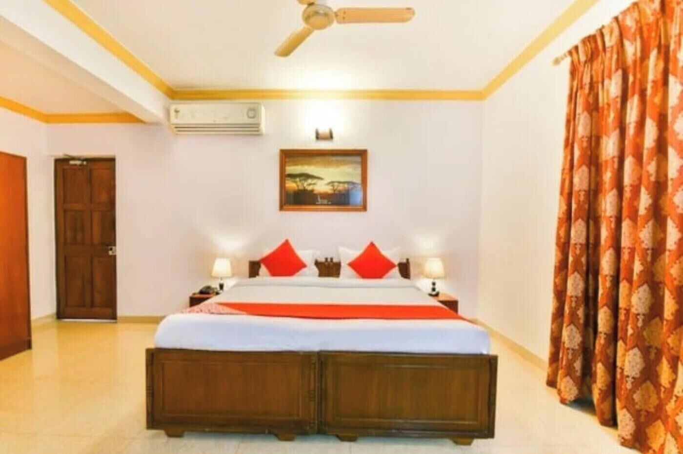 A hotel room with a bed, two bedside tables, a ceiling fan, an air conditioning unit, a painting on the wall, and a patterned curtain.