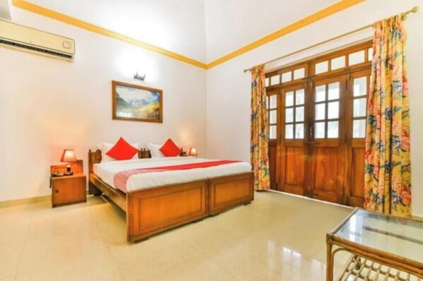 A bedroom with a wooden bed, a nightstand, a bedside lamp, a window with curtains, and a door. - Joy Guest House (Calangute)