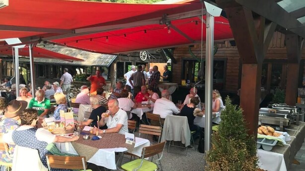 An outdoor dining area with tables and chairs under red canopies, surrounded by wooden structures and greenery. - Waldrestaurant Vida  (Kapfenberg)