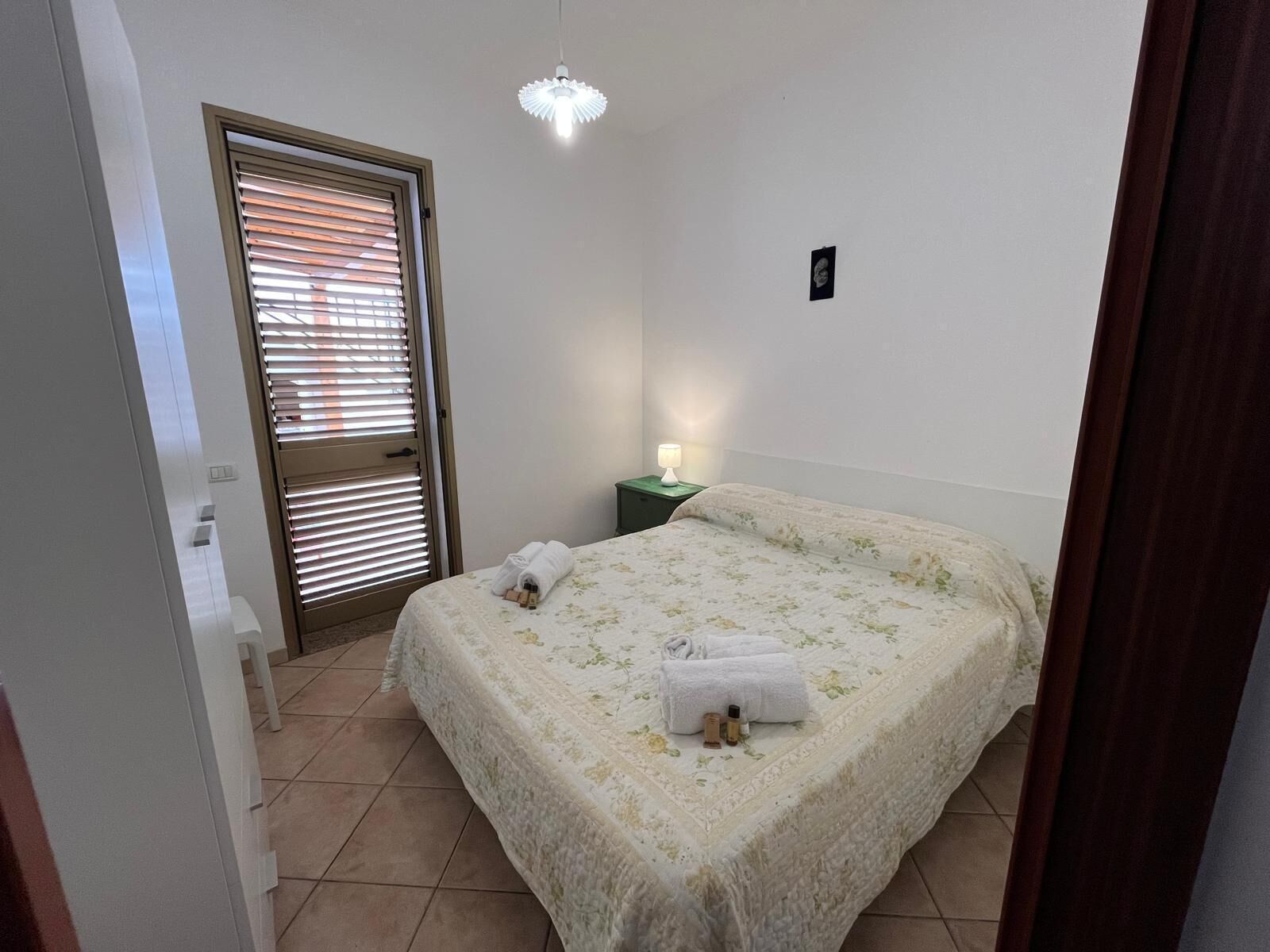 A bedroom with a bed, a nightstand, a lamp, and a door with blinds.