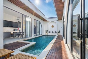 A modern indoor pool area with a wooden deck, a dining area with a table and chairs, and a kitchen with a TV. - Casa Santisya 4 (Munggu)