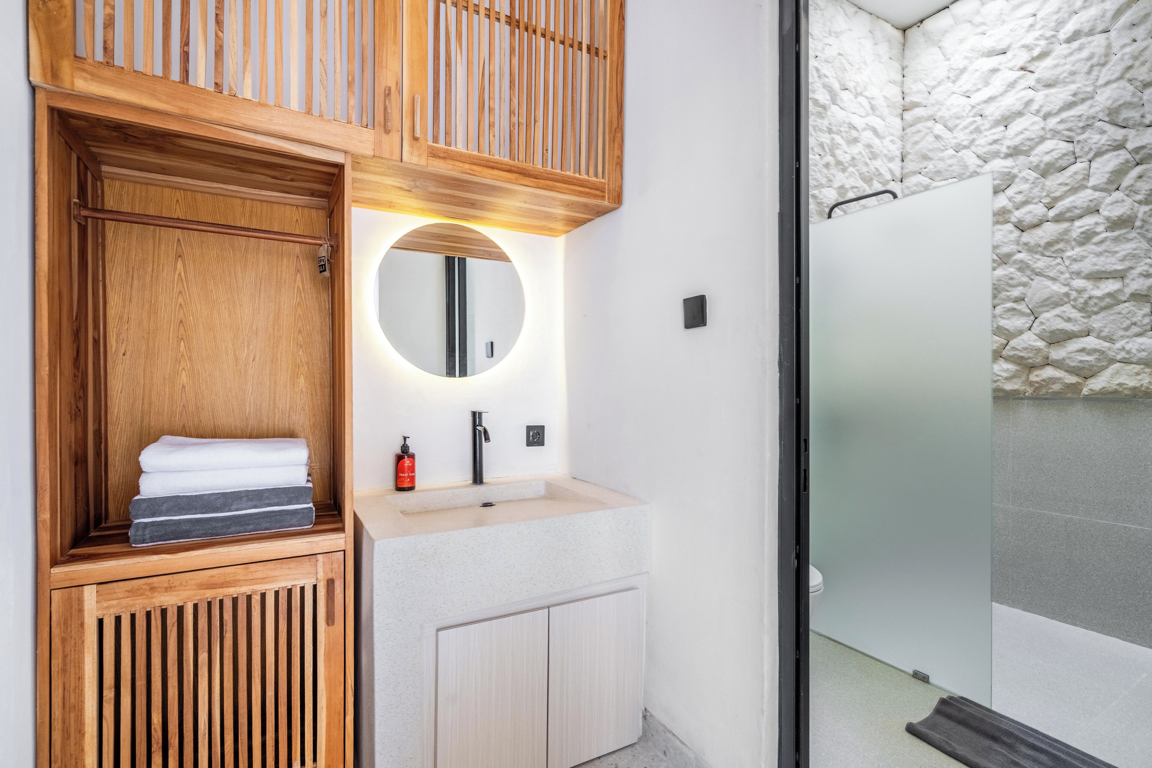 A modern bathroom with a wooden vanity, a round mirror, and a stone wall.