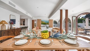 Comedor con mesa de madera dispuesta para seis comensales, un televisor de pantalla grande al fondo y vistas al exterior a través de amplias ventanas.