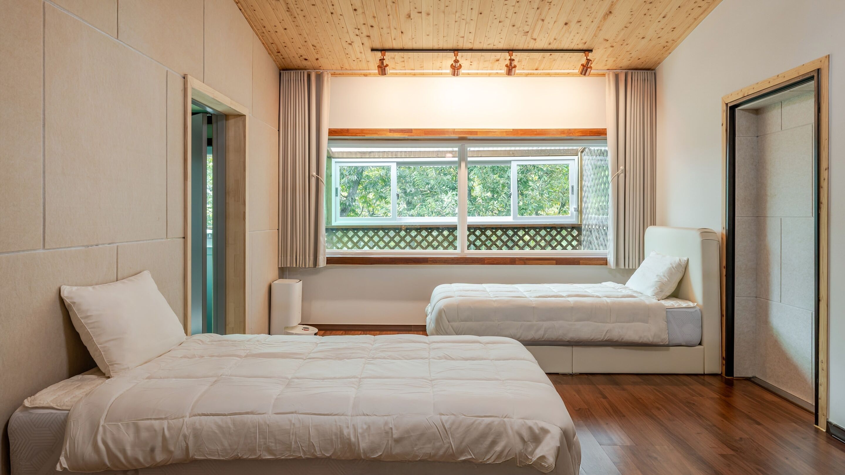 A hotel room with two beds, a window with a view of greenery, wooden flooring, and a wooden ceiling.