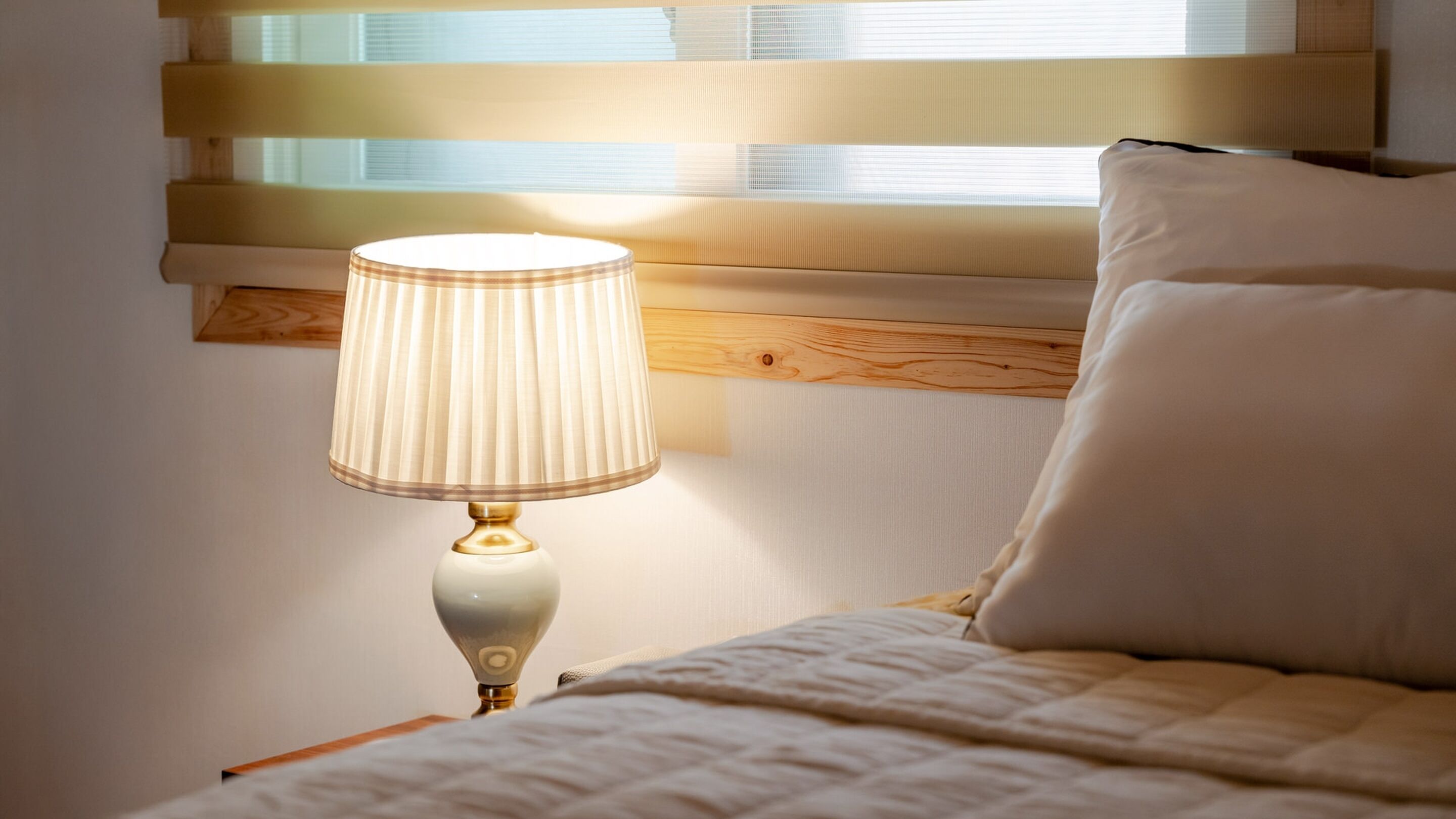 A bed with a white quilt, a bedside lamp with a white shade, and a window with blinds.