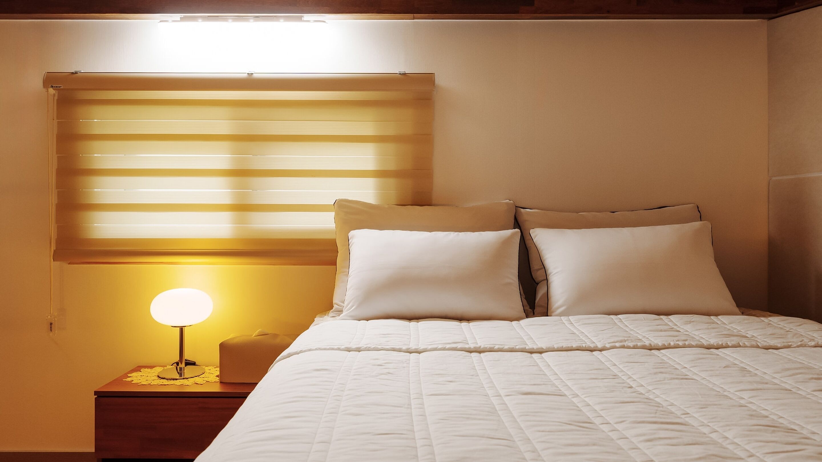A bedroom with a bed, pillows, a bedside table with a lamp, and a window with blinds.