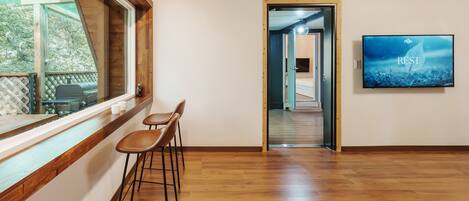 A room with a wooden floor, a large window, two bar stools, a television, and a door leading to another room.