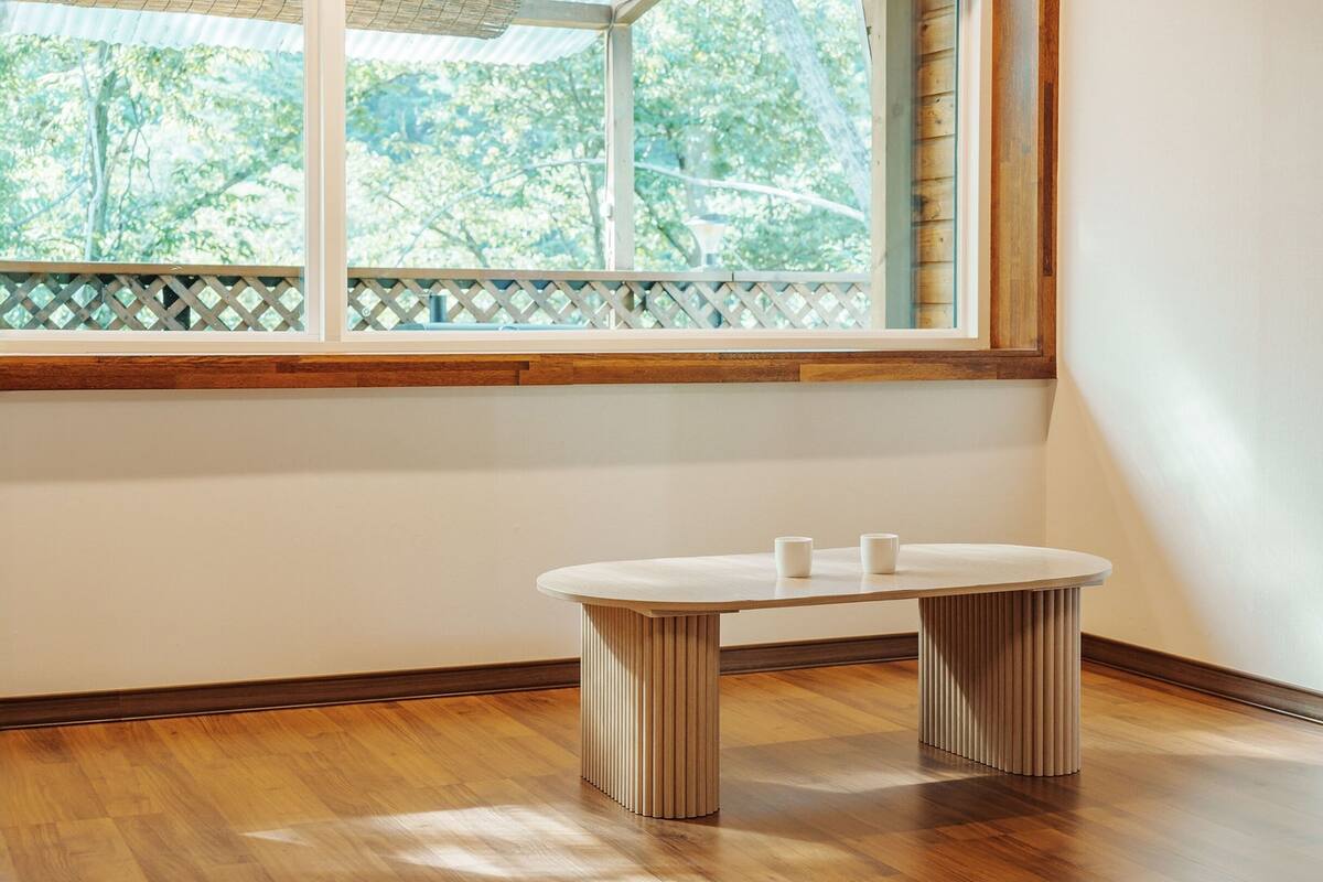 Una habitación con piso de madera, ventana con vista a árboles y una mesa redonda con dos tazas blancas.