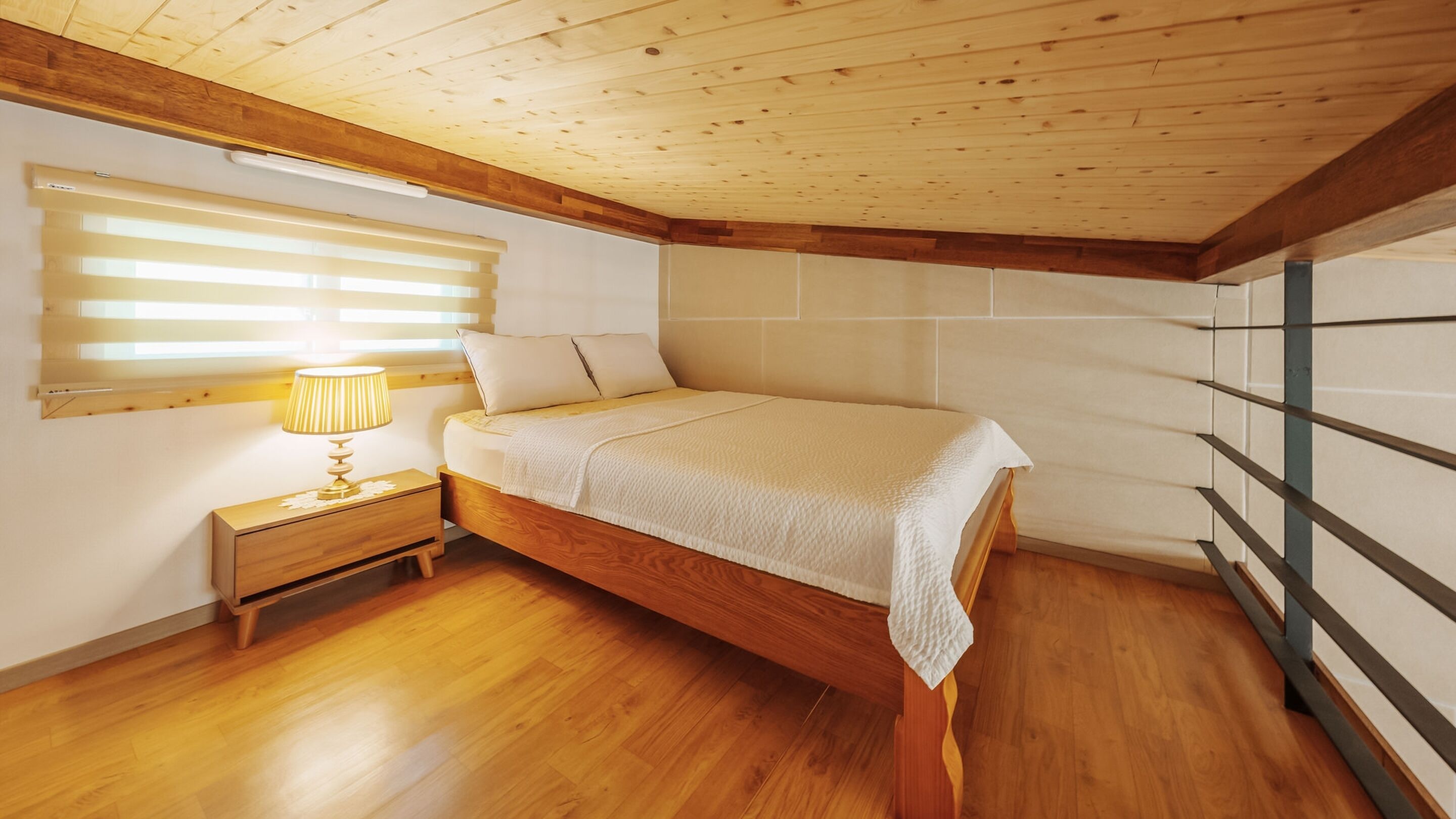 A bedroom with a wooden bed, a nightstand with a lamp, wooden flooring, and a sloped ceiling.