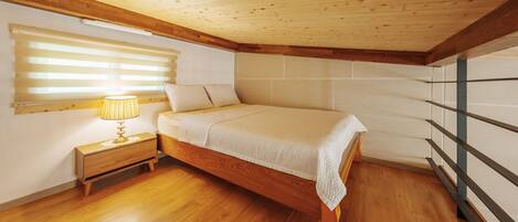 A bedroom with a wooden bed, a nightstand with a lamp, wooden flooring, and a sloped ceiling.