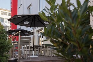 Outdoor seating with umbrellas, a building with a red and white facade, and a plant in the foreground. - Trip Inn Goethe Hotel Messe Frankfurt (Frankfurt)
