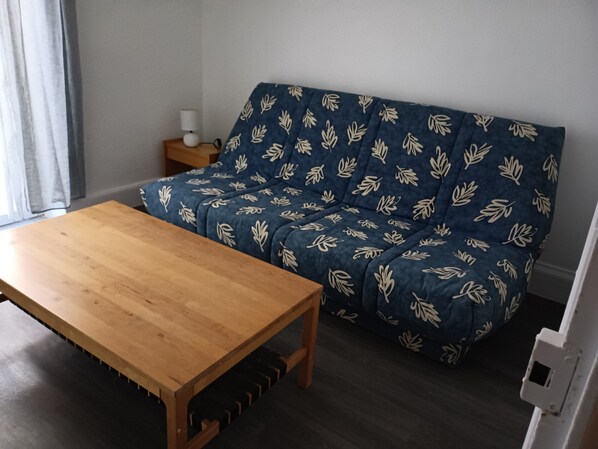 A room with a wooden table, a blue sofa with leaf patterns, a small bedside table with a lamp, and a dark wooden floor. - LUXURY APARTMENT (Vichy)
