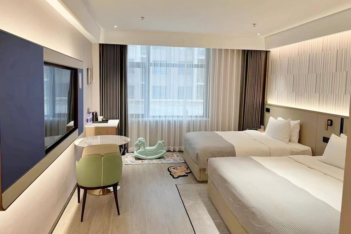 Quarto de hotel moderno com uma cama grande, uma cama menor, uma escrivaninha com cadeira, televisão e uma janela ampla com cortinas.