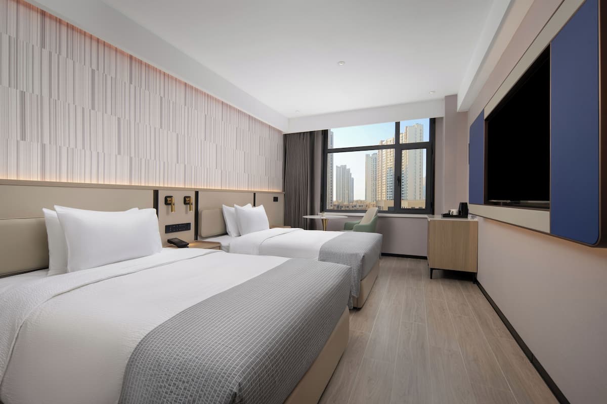 Quarto de hotel com duas camas, uma escrivaninha, uma cadeira, uma televisão e janela com vista para a cidade.