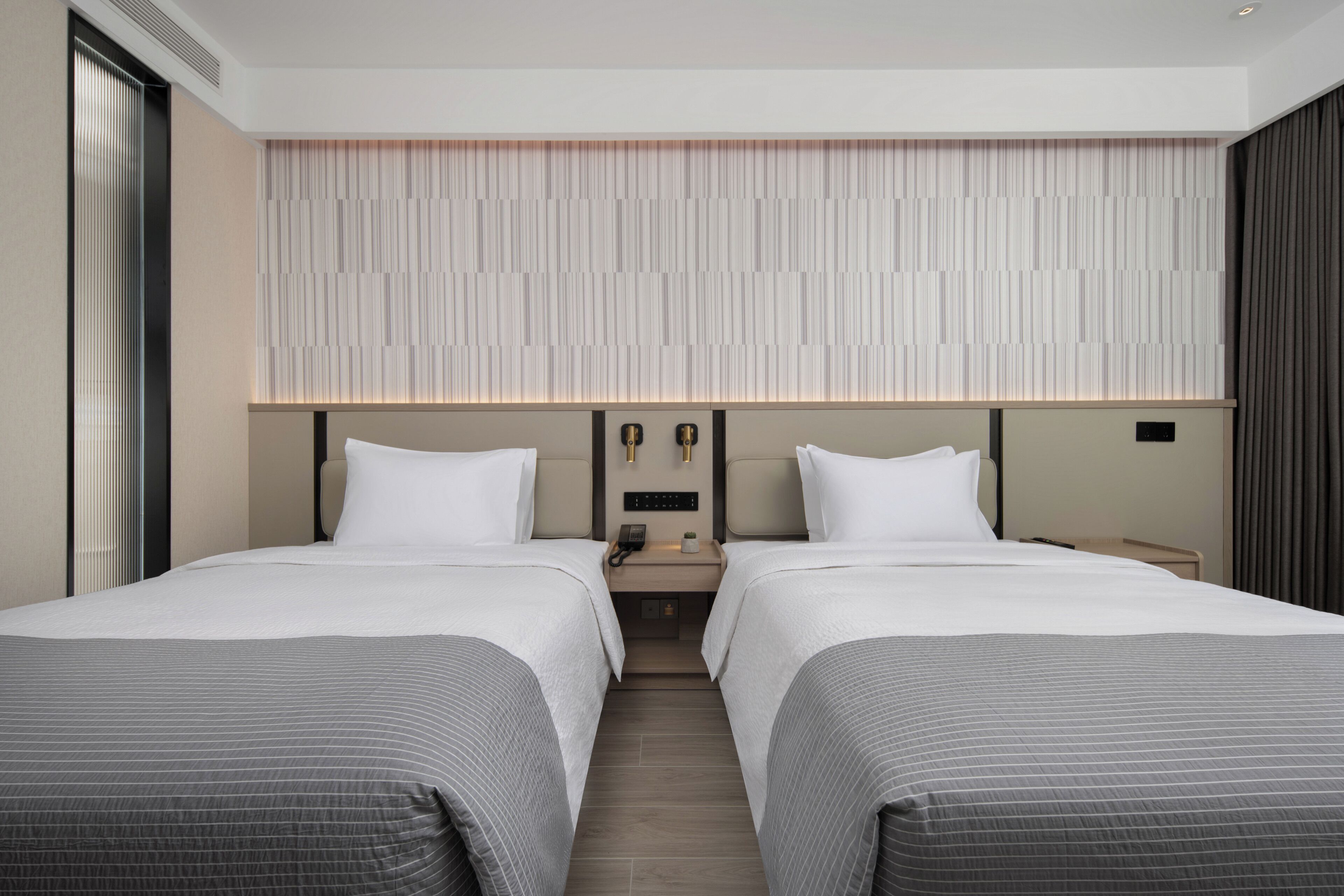 Une chambre d’hôtel moderne avec deux lits, une grande fenêtre, un panneau de commande fixé au mur et une table de chevet avec une lampe.