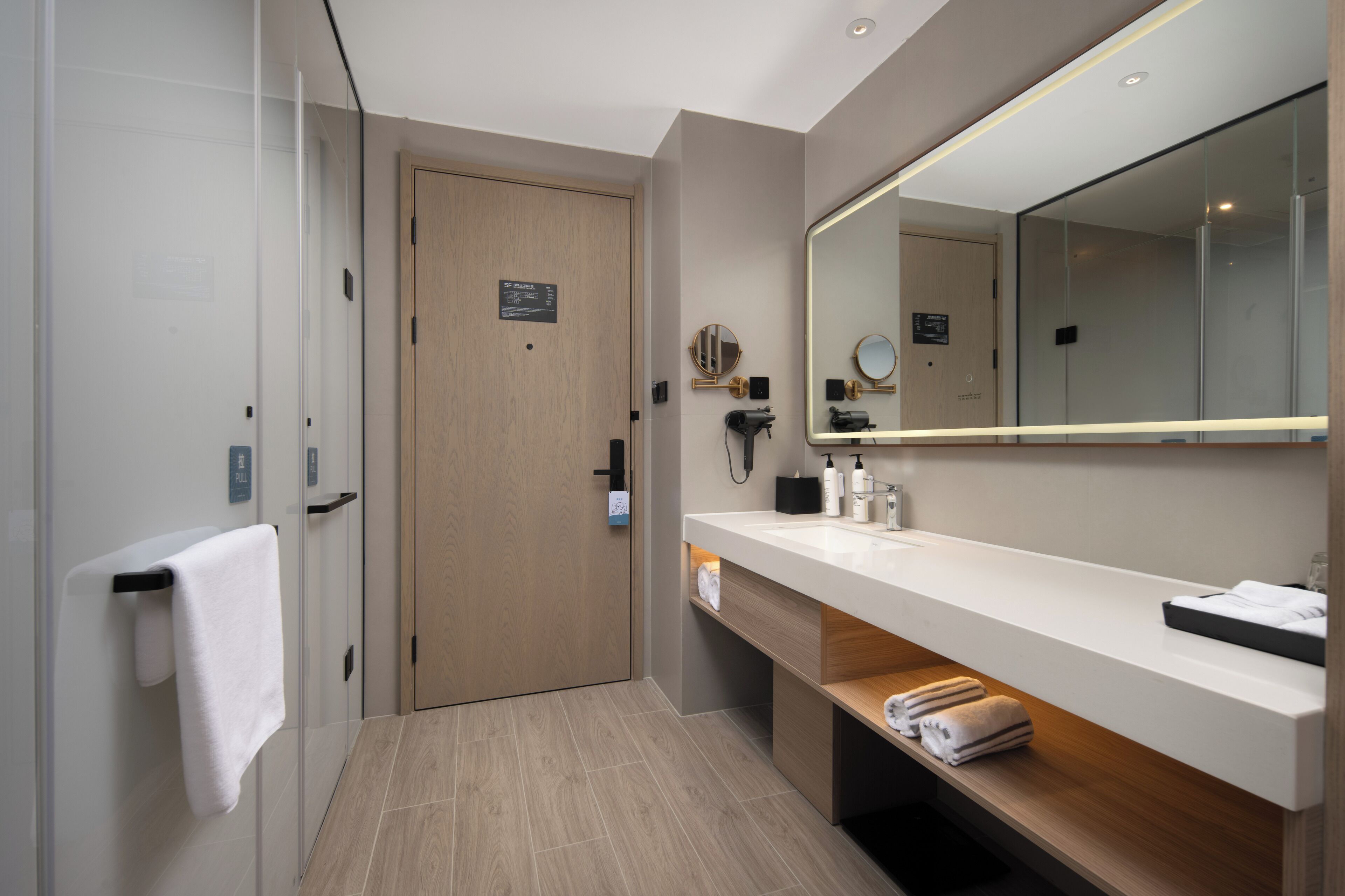 A modern bathroom with a glass shower enclosure, a wooden door, a large mirror, and a white countertop with toiletries.