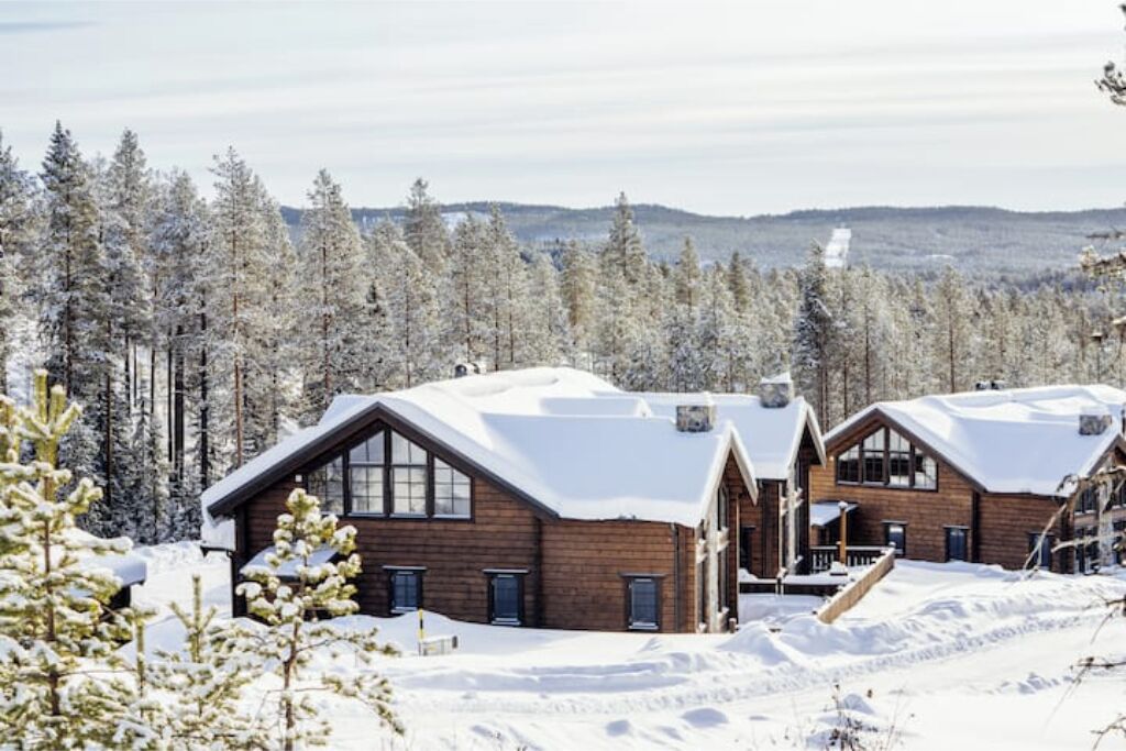 Una cabaña de madera con techo inclinado, rodeada de árboles cubiertos de nieve y un paisaje montañoso.
