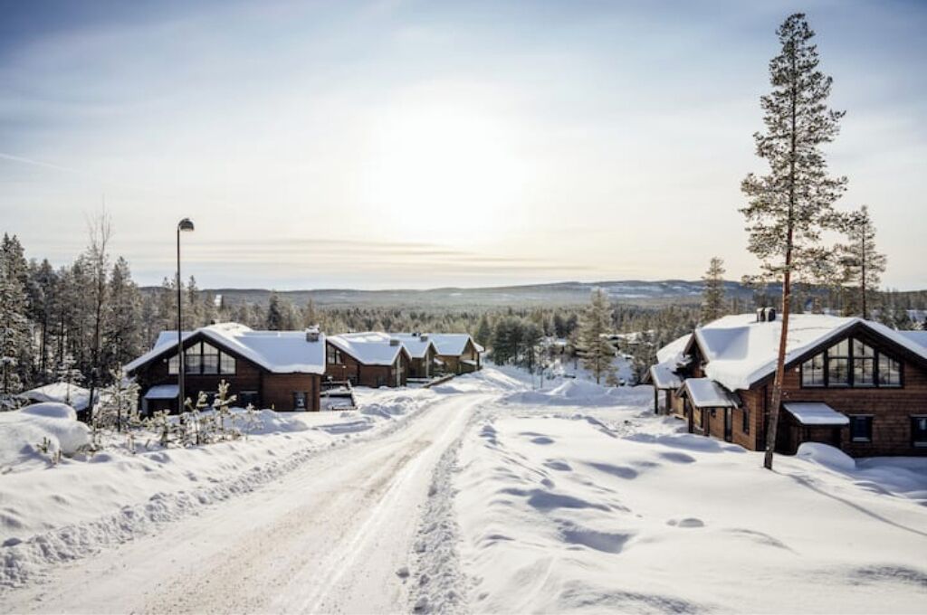 Une route enneigée mène à une rangée de chalets en bois, d’où l’on a une vue dégagée sur la forêt environnante et les collines lointaines.