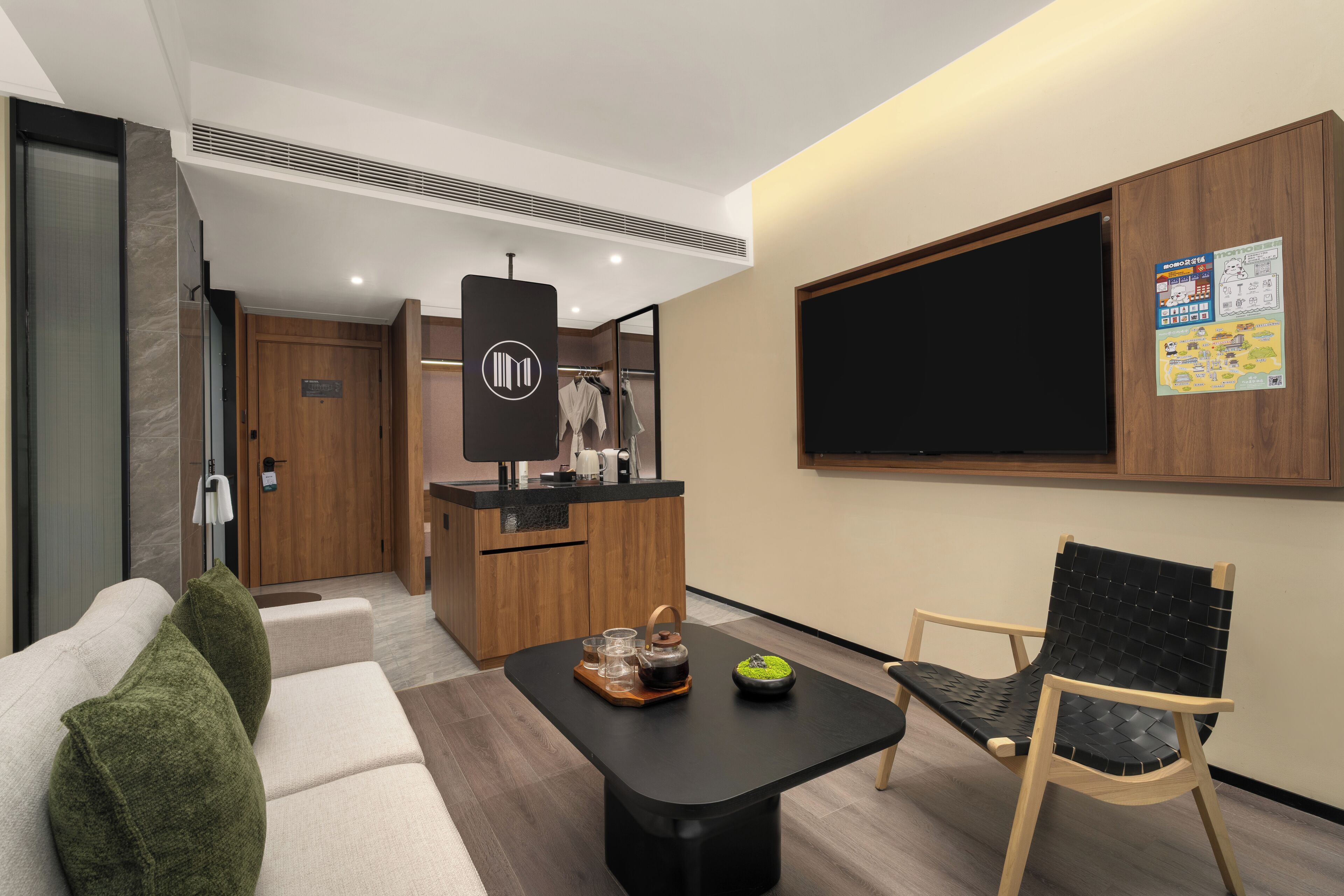 A modern hotel room with a sofa, a dining table with a kettle and a bowl, a wooden chair, a kitchen area, and a large window.