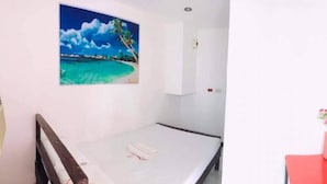 A small, single-bed room with a painting of a beach scene, a small table with a vase of flowers, and a red bench. - WJV INN CARCAR (Carcar)