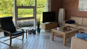 A living room with a TV, sofa, armchair, and a large window with a view of greenery. - Lovely holiday home near beach and nature (Karrebæksminde)