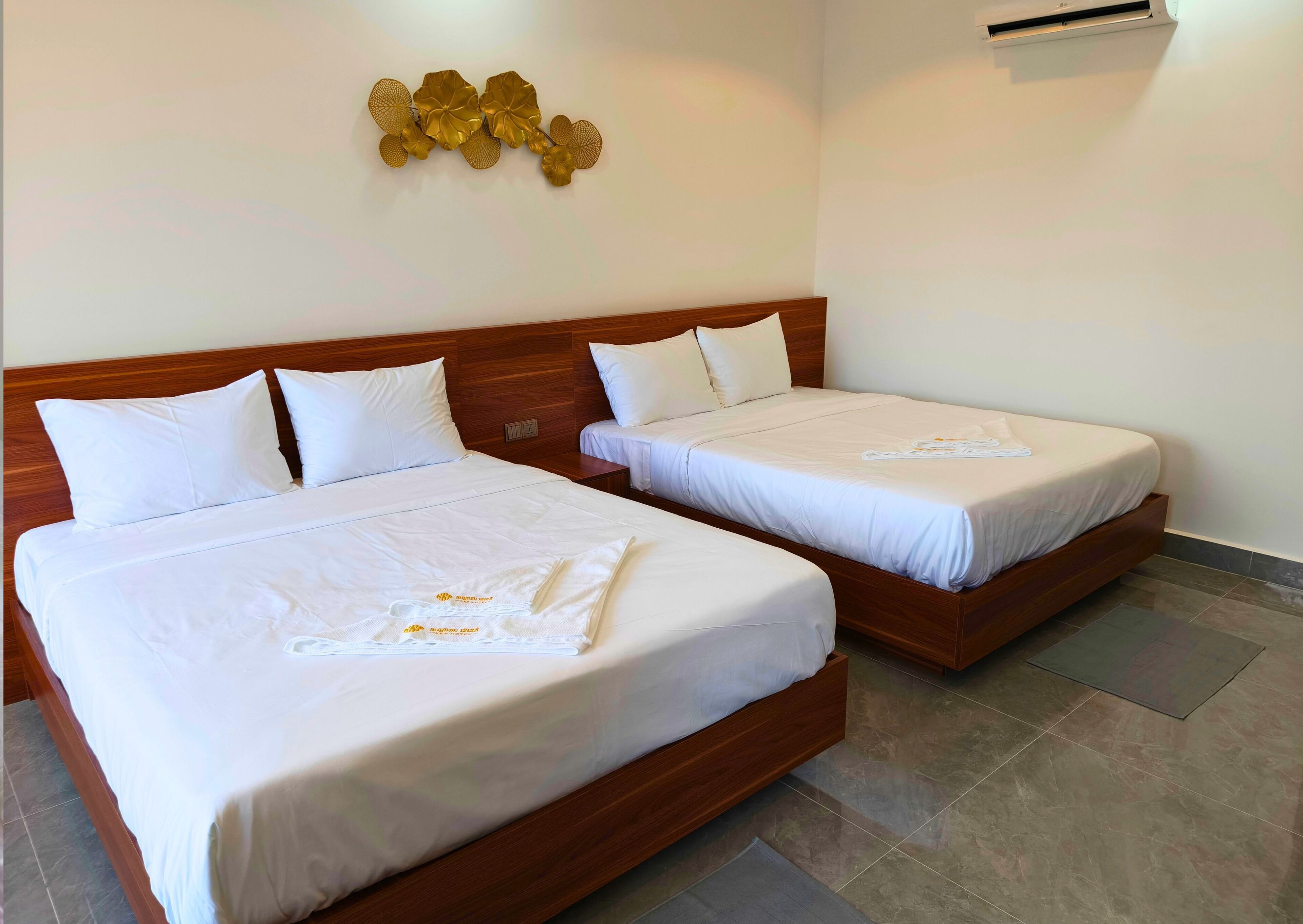 Habitación de hotel con dos camas individuales, ropa de cama blanca, cabecero de madera, aire acondicionado de pared y un adorno dorado en la pared.