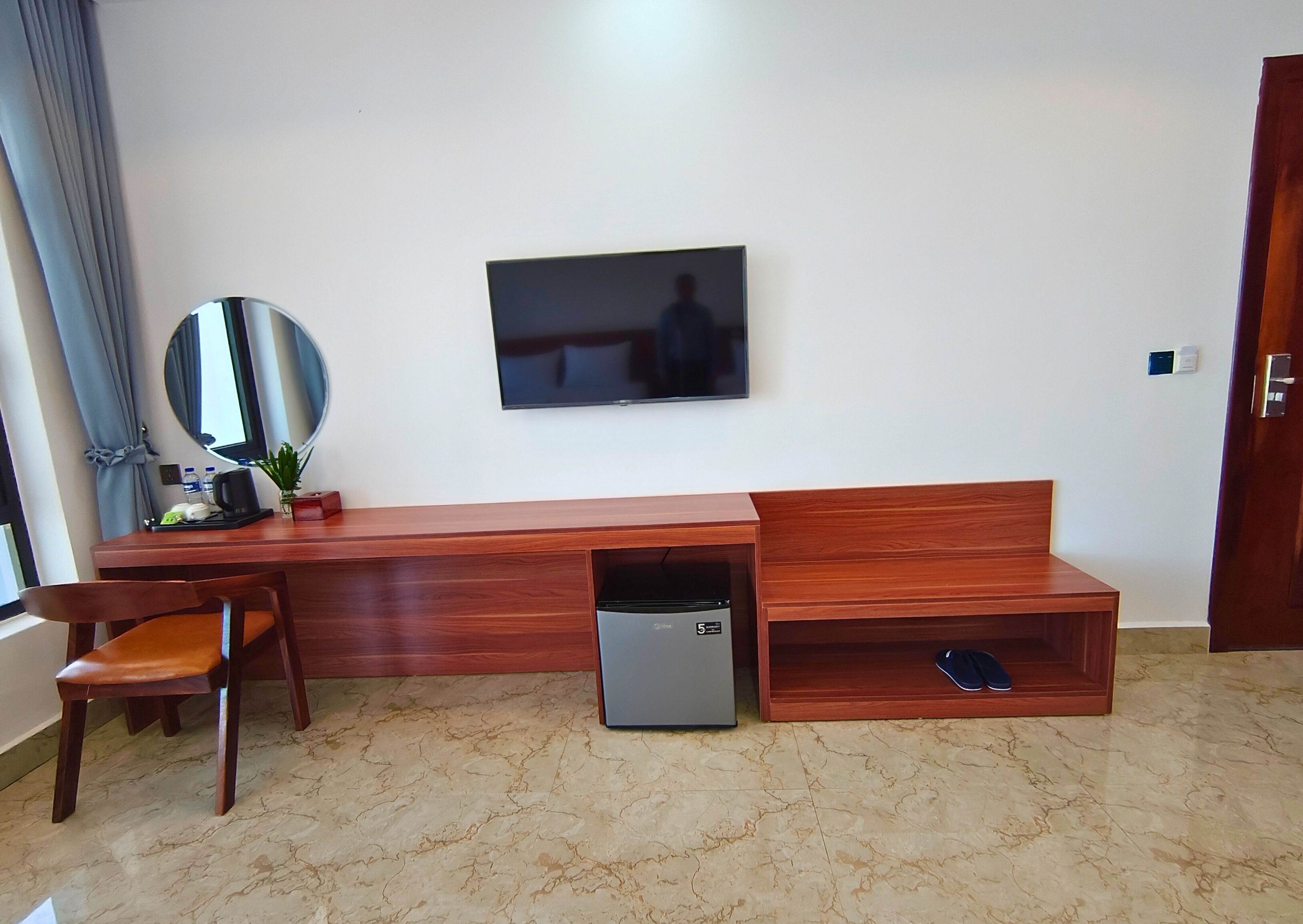 A hotel room with a wooden desk, a chair, a television mounted on the wall, a small refrigerator, and a door.