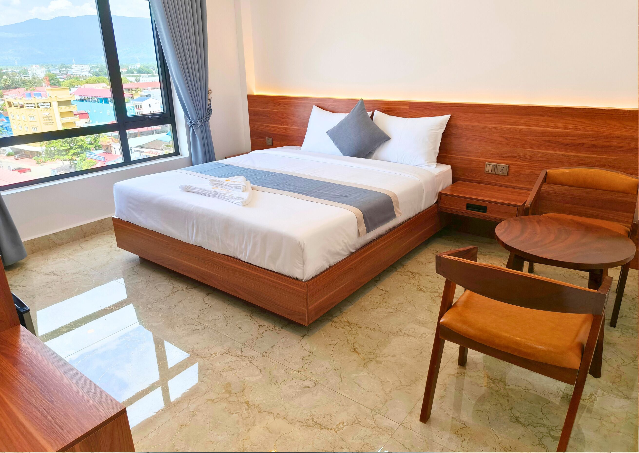 Une chambre d’hôtel avec un grand lit, une tête de lit en bois, une petite table, une chaise et une vue sur les bâtiments par la fenêtre.