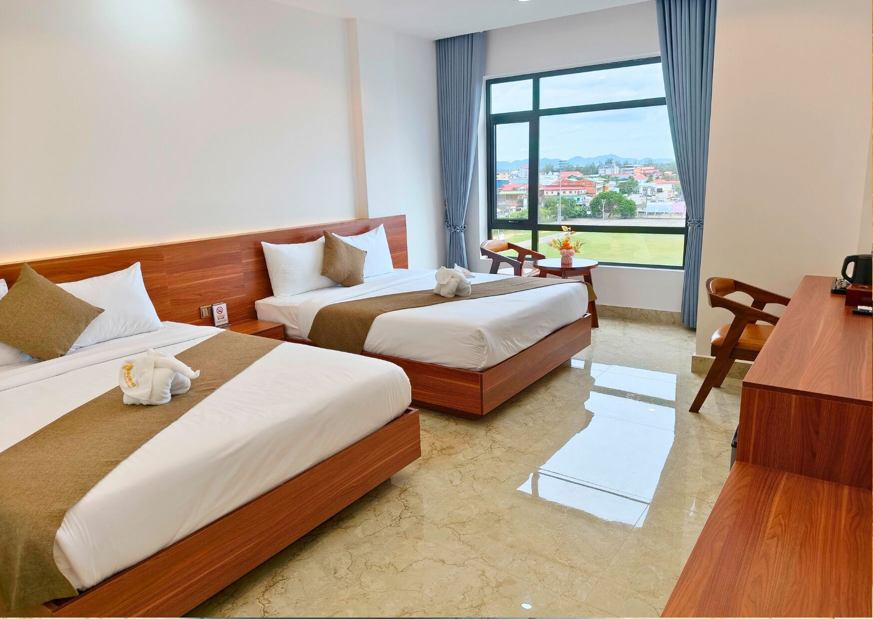 A hotel room with two beds, a desk, and a view of a cityscape through large windows.