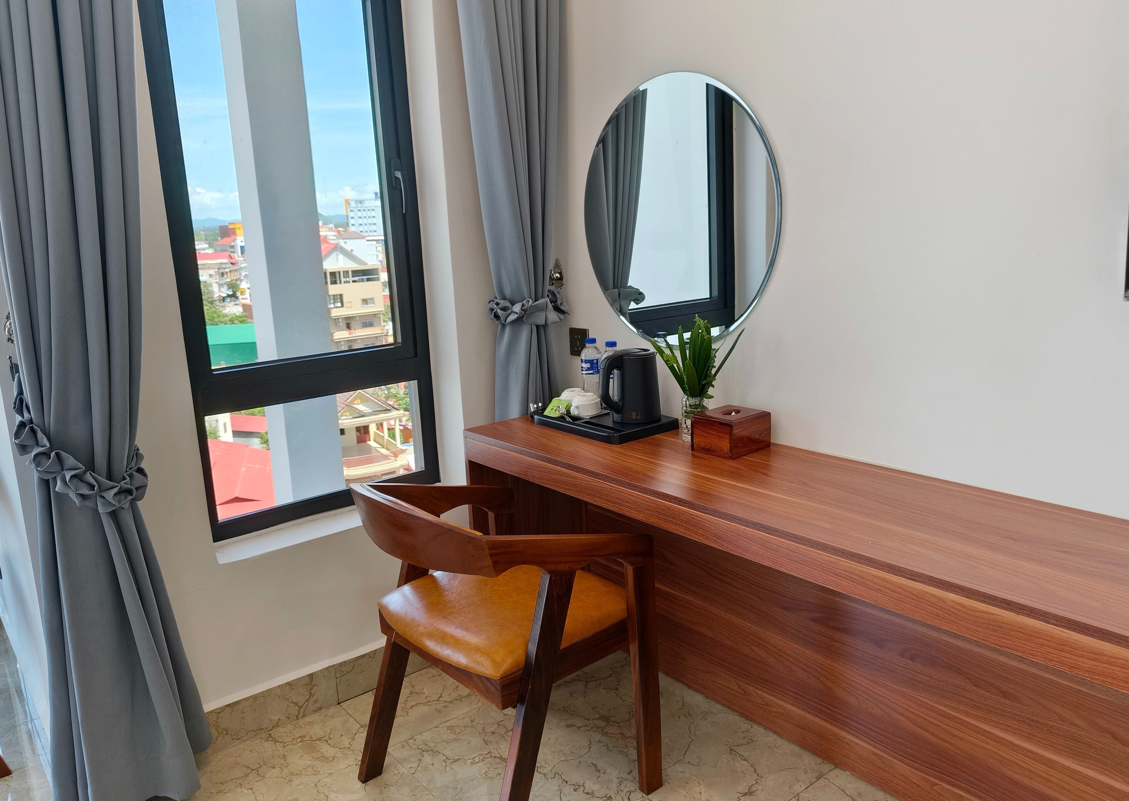 A hotel room with a wooden desk, a chair, a large mirror, and a window with curtains.