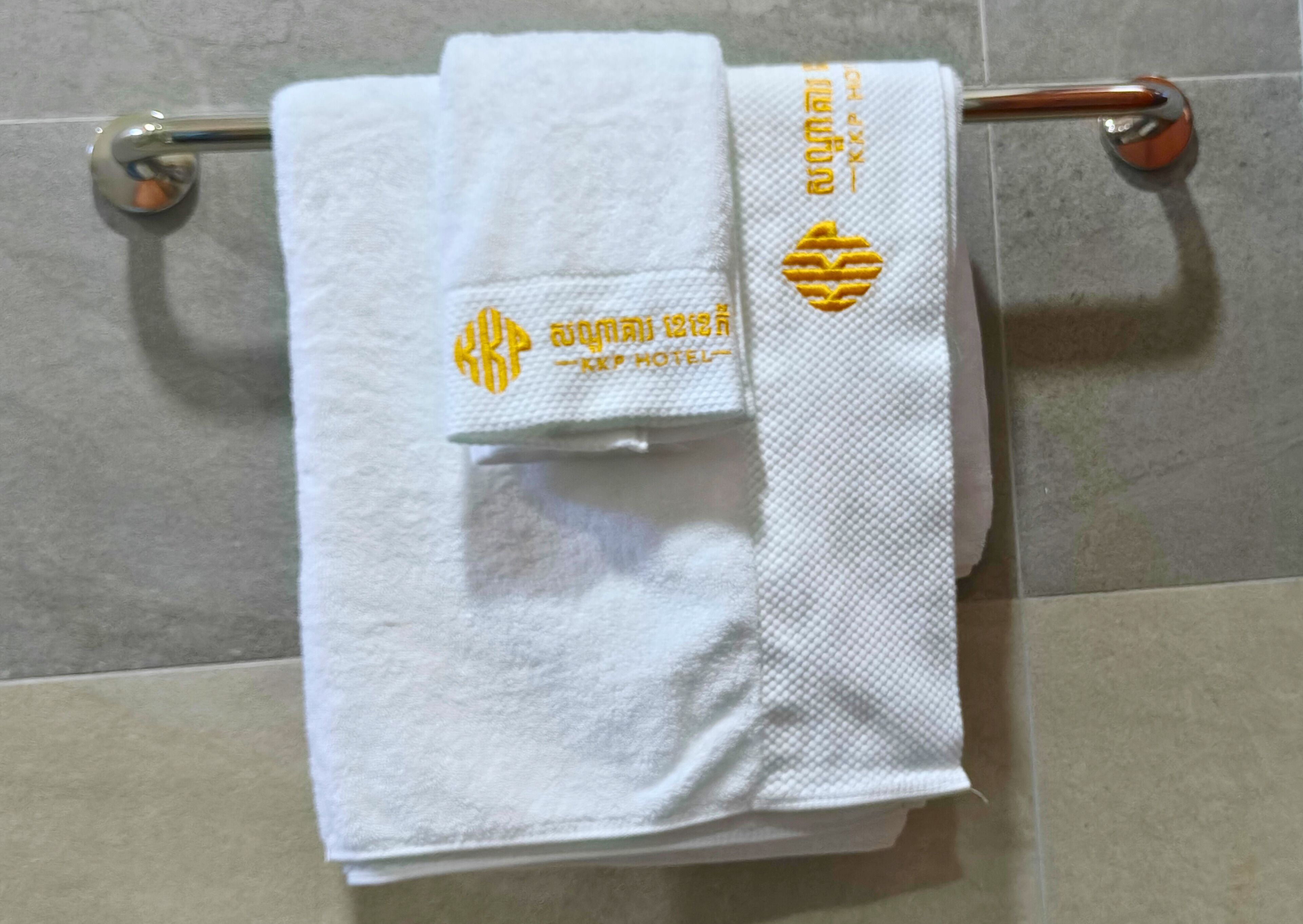 A white towel with a logo and text, hanging on a metal towel rack against a tiled wall.