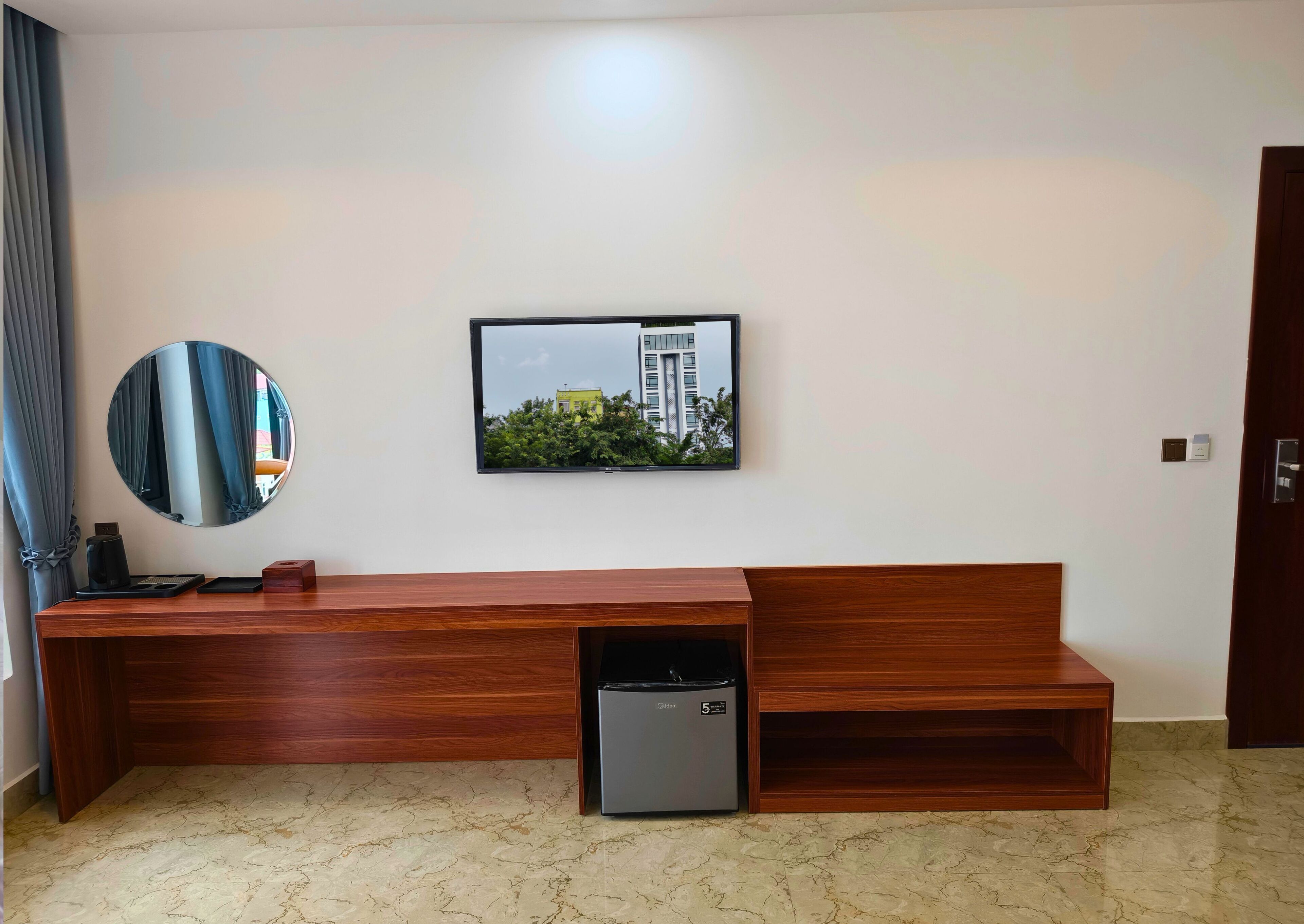 A modern hotel room with a wooden console table, a small refrigerator, a round mirror, and a framed picture of a building.