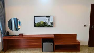 A modern hotel room with a wooden console table, a small refrigerator, a round mirror, and a framed picture of a building.