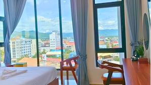 Une chambre d’hôtel avec un lit, un bureau et une chaise. Une grande fenêtre offre une vue sur les bâtiments et les montagnes.