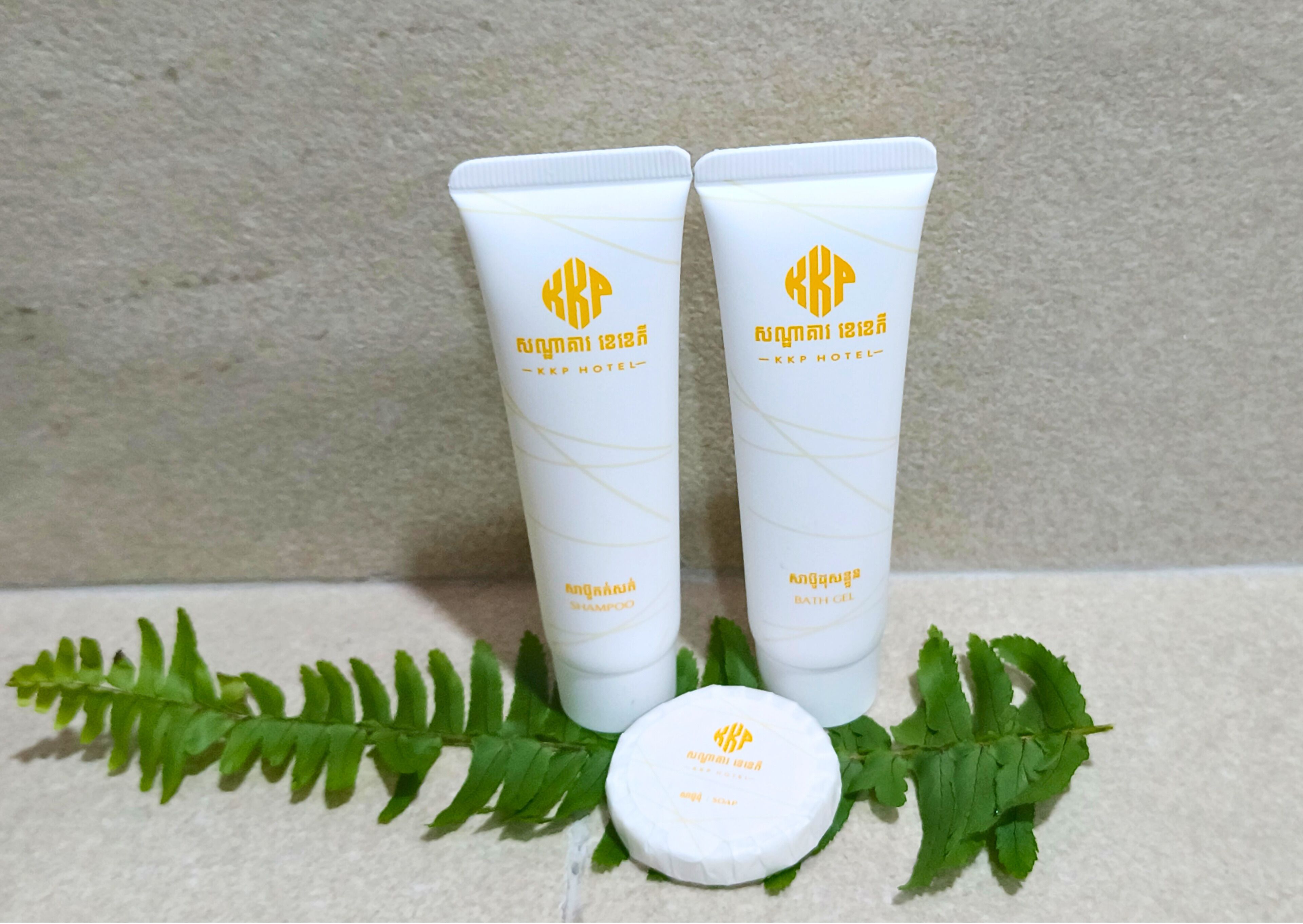 Two white tubes with yellow logos and text, one labeled 'shampoo' and the other 'bath gel', placed on a surface with a green fern leaf and a white circular object.