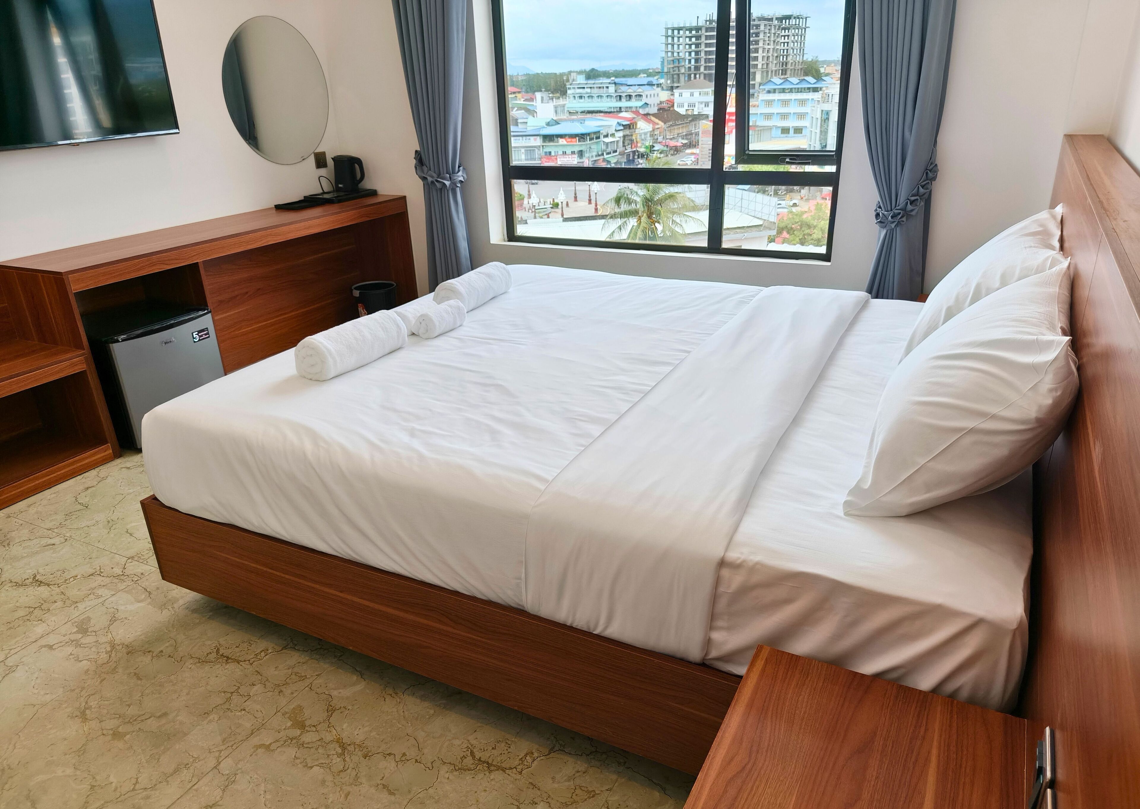 A hotel room with a large bed, wooden headboard, a small desk with a mirror, a coffee maker, and a view of the city through the window.