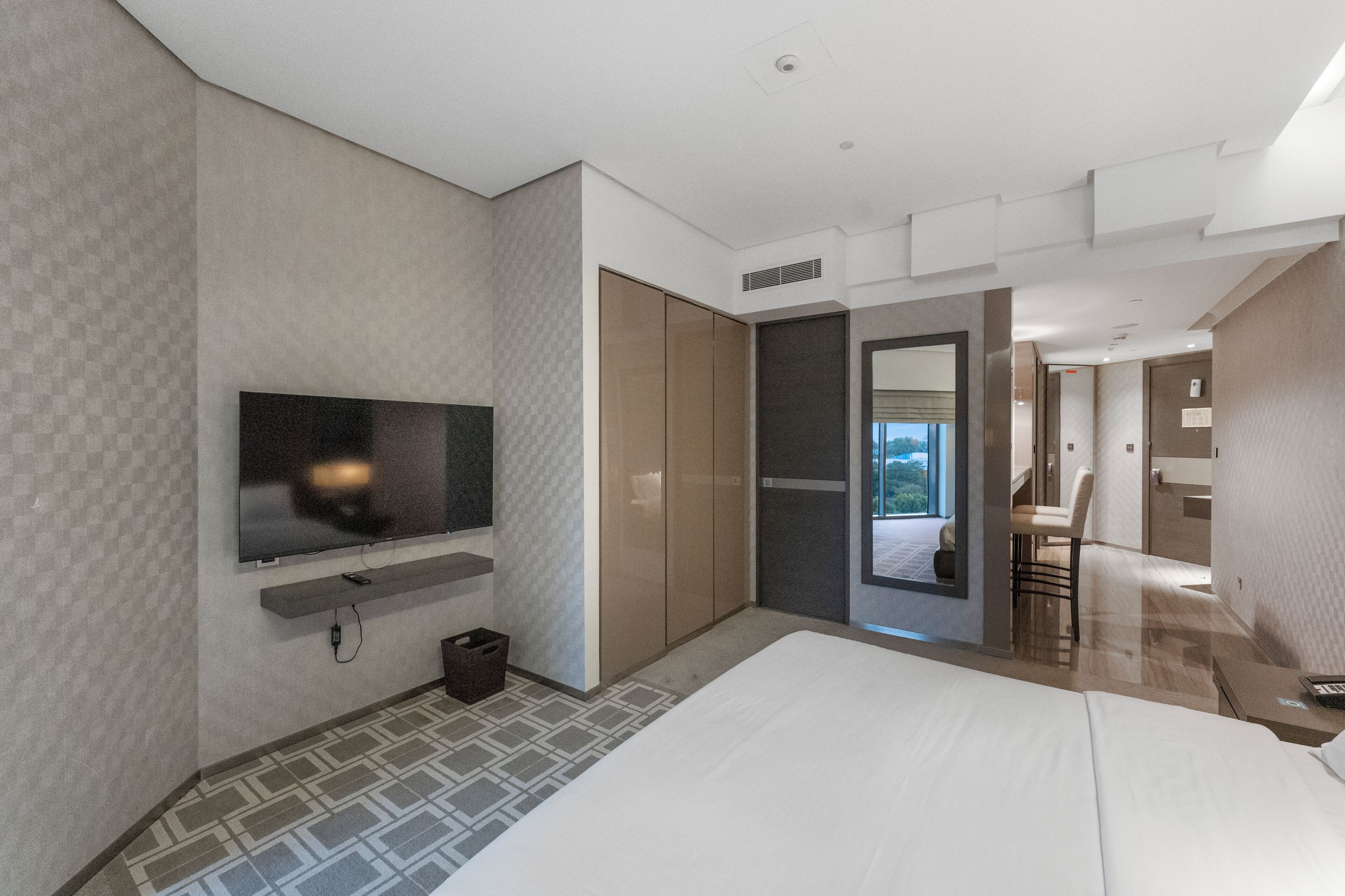 A modern hotel room with a large bed, a flat-screen TV mounted on the wall, a small seating area with a chair, and a view of the outdoors through a window.