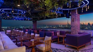A rooftop lounge with seating and tables, a city skyline view, and hanging lights.