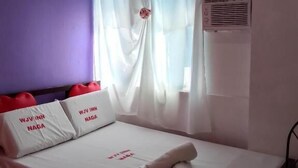 A hotel room with a bed, white bedding, red heart-shaped pillows, a window with sheer curtains, and an air conditioning unit. - WJV INN NAGA (Naga City)