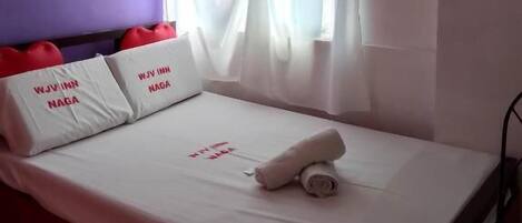 Habitación de hotel con cama, ropa de cama blanca, almohadas rojas con forma de corazón, ventana con cortinas traslúcidas y un equipo de aire acondicionado.
