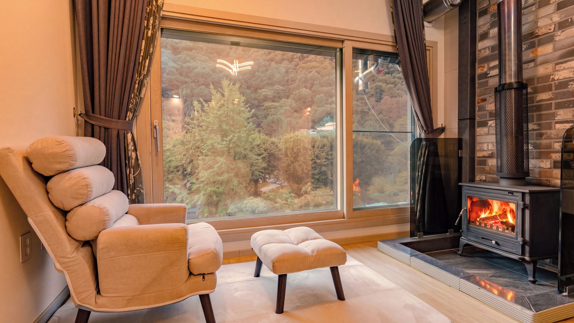 A cozy living room with a large window, a comfortable armchair, a small ottoman, and a wood-burning stove.