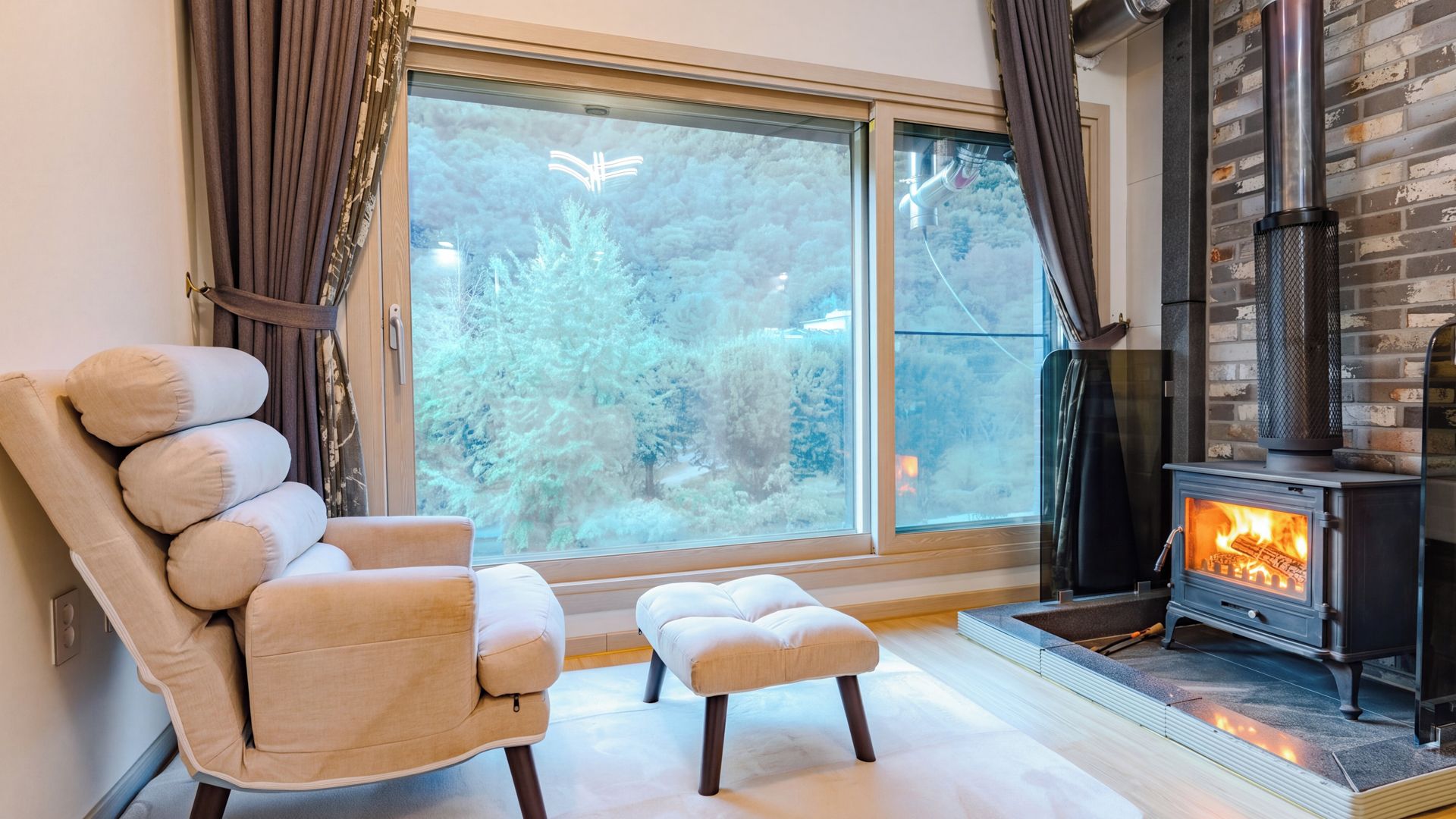 Una habitación acogedora con un ventanal grande, un sillón cómodo, un pequeño reposapiés y una estufa de leña.