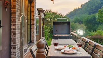 Un área de comedor al aire libre con una parrilla, mesa, sillas y una vista panorámica de una ladera boscosa.